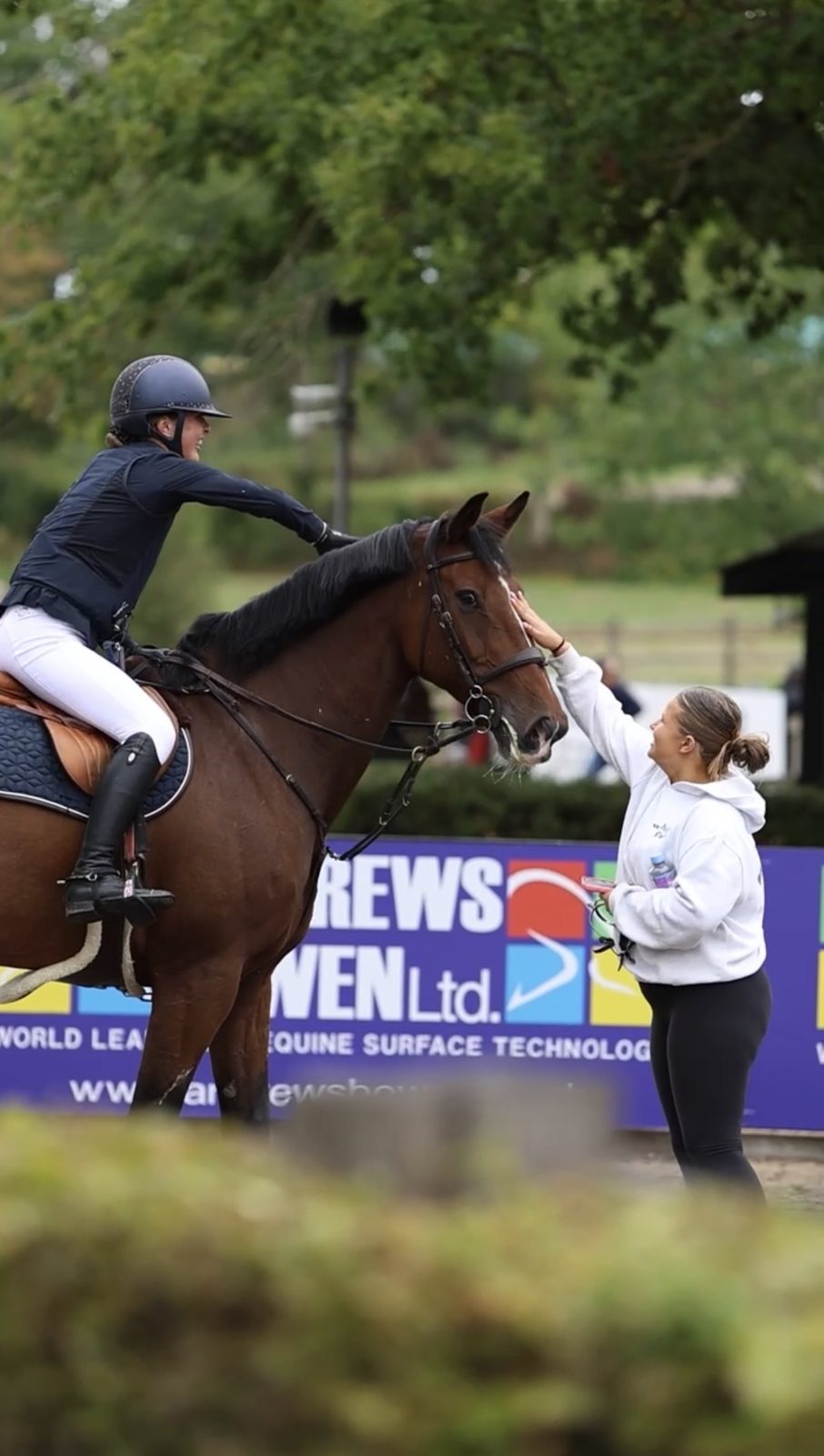 A smiling rider and a supporter both patting a bay horse after its round